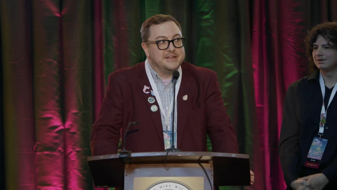 Brent Fisher speaking into a microphone at the Ringo Awards podium, in a maroon jacket covered in enamel pins, against a stage curtain lit red and green.