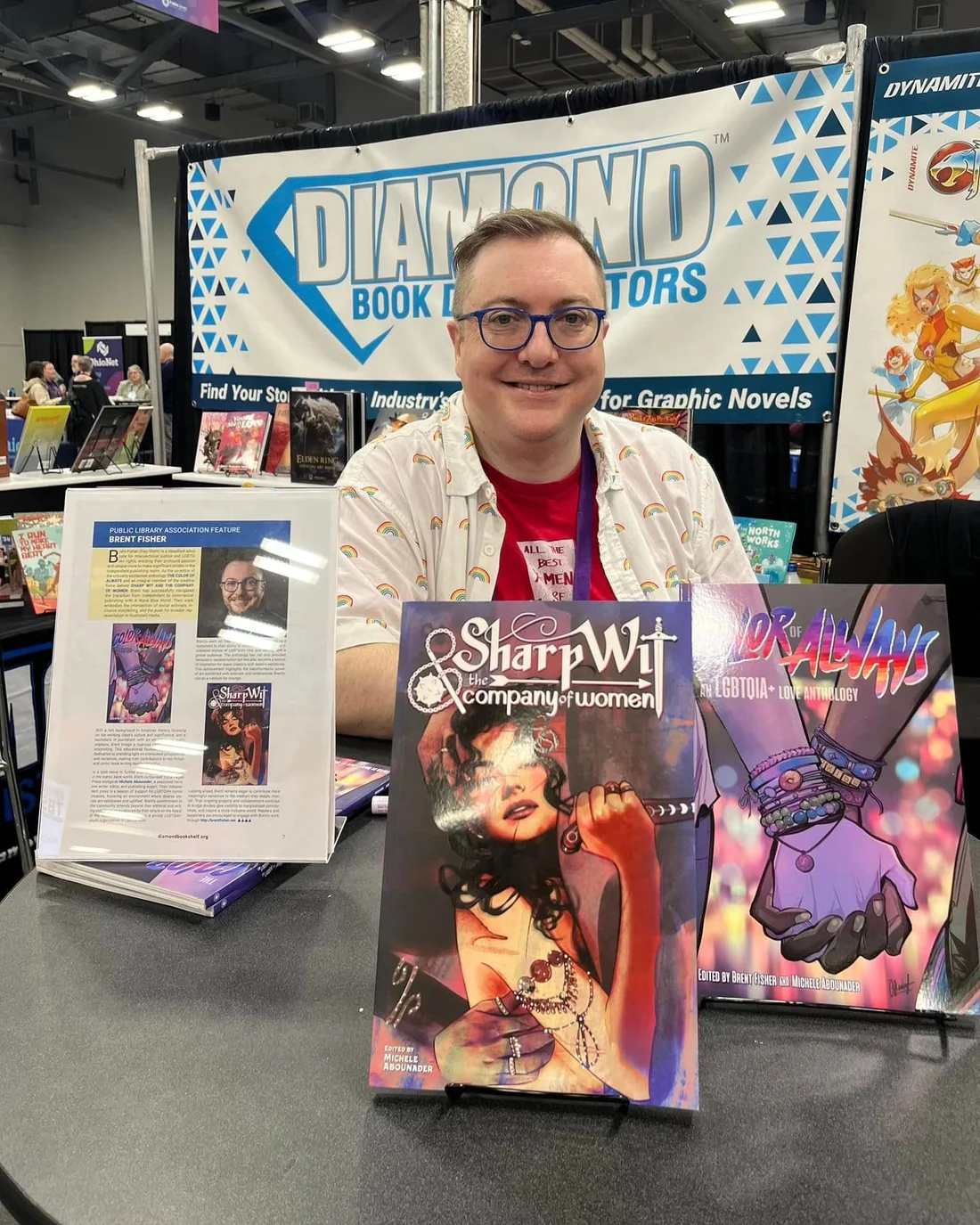 Brent Fisher seated at a convention table beneath a Diamond Book Distributors banner, with copies of Sharp Wit & the Company of Women and The Color of Always standing on the table in front of them.